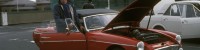 Photograph of a man stepping out of his red MG midget car. The bonnet is up.