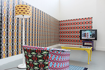 A shot of an installation with a brightly patterned armchair and standard light, with patterned wall paper on the walls and a TV